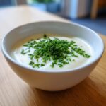Nahaufnahme einer cremigen Blumenkohlsuppe in einer weißen Schüssel, garniert mit viel frischem Schnittlauch.