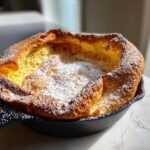 Ein goldbraun gebackener Dutch Baby zum Muttertag, bestäubt mit Puderzucker, in einer gusseisernen Pfanne.