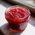 Nahaufnahme von leuchtend roter Erdbeer Rhabarber Marmelade in einem kleinen Glas, beleuchtet von Sonnenlicht.