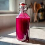 Eine Glasflasche gefüllt mit leuchtend pinkem Rhabarber-Sirup steht auf einer hellen Arbeitsplatte im Sonnenlicht.