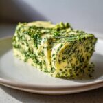 Ein Block selbstgemachte Bärlauchbutter mit viel grünen Kräutern auf einem hellen Teller.