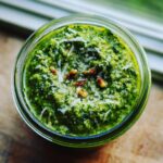 Aufnahme von oben in ein Glas mit leuchtend grünem Bärlauchpesto, garniert mit geriebenem Käse und Pinienkernen.