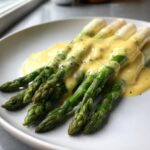 Nahaufnahme von grünem Spargel, bedeckt mit cremiger Sauce Hollandaise und schwarzem Pfeffer.