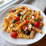 Nahaufnahme von Italienischer Nudelsalat mit Fusilli, Kirschtomaten, Feta und schwarzen Oliven auf einem weißen Teller.