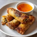 Vier knusprige, goldbraun frittierte Spargel-Frühlingsrollen auf einem Teller mit einem kleinen Schälchen süß-saurer Dip.