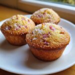 Drei goldbraune Rhabarber-Muffins mit grobem Zuckerguss auf einem weißen Teller.