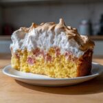 Ein dickes Stück Rhabarberkuchen mit Baiser, das auf einem weißen Teller liegt und viel saftigen Rhabarber zeigt.