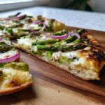 Nahaufnahme eines angeschnittenen Stücks Spargel-Flammkuchen mit grünem Spargel und roten Zwiebelringen auf einem Holzbrett.