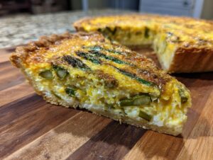 Nahaufnahme eines Stücks Spargelquiche mit grünem Spargel und goldbrauner Kruste auf einem Holzbrett.