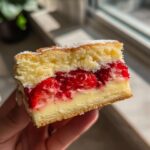 Nahaufnahme eines Stücks Erdbeerkuchen mit Pudding, gehalten in der Hand, mit viel saftigen Erdbeeren.