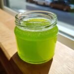 Nahaufnahme von leuchtend grünem Waldmeistergelee in einem kleinen Glas, das im Sonnenlicht steht.