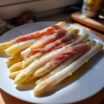 Nahaufnahme von gekochtem Weißer Spargel mit Schinken, angerichtet auf einem weißen Teller mit etwas Öl.