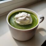 Nahaufnahme einer leuchtend grünen Bärlauchschaumsuppe in einer weißen Tasse, gekrönt mit einem großen Klecks Schlagsahne.