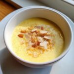 Nahaufnahme einer Schüssel mit gelber, cremiger Pfingstmilch, garniert mit Mandelblättchen und ganzen Mandeln.