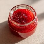 Nahaufnahme einer offenen Glas mit leuchtend roter Erdbeer-Rhabarber-Marmelade, die im Sonnenlicht glänzt.
