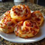 Nahaufnahme von fünf goldbraunen, gebackenen Pizzabrötchen mit geschmolzenem Käse und Salami auf einem weißen Teller.