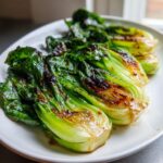 Nahaufnahme von mehreren Stücken gebratenem Pak Choi, glasiert mit einer dunklen Soße, auf einem weißen Teller.