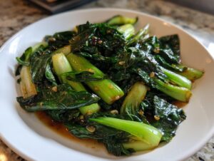 Nahaufnahme von saftigem, gebratenem Pak Choi in der Pfanne mit dunkler Soße auf einem weißen Teller.