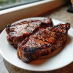 Nahaufnahme von zwei saftigen, gegrillten Nackensteaks mit dunklen Grillstreifen auf einem weißen Teller.
