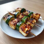 Nahaufnahme von zwei gegrillten Halloumi-Zucchini-Spießen mit schön gebräuntem Käse auf einem hellen Teller.