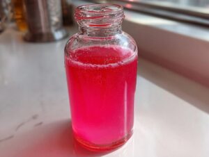 Nahaufnahme von intensiv pinkem Rhabarbersirup in einer kleinen Glasflasche, die auf einer hellen Oberfläche steht.