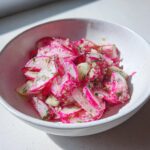 Nahaufnahme eines knackigen Radieschensalat mit rosa Scheiben und Kräutern in einer weißen Schüssel.