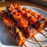 Nahaufnahme von gegrillten Currywurst-Spieße, übergossen mit leuchtend oranger Currysoße auf einem weißen Teller.