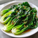 Nahaufnahme von leuchtend grünem Pak Choi gebraten, bedeckt mit einer würzigen Knoblauch-Chili-Soße auf einem weißen Teller.