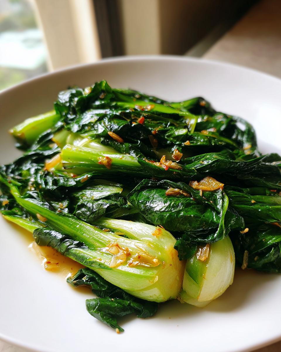 Nahaufnahme von leuchtend grünem, gebratenem Pak Choi mit Knoblauchstückchen auf einem weißen Teller.