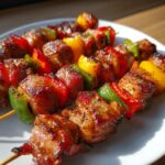 Mehrere gegrillte Schweinefilet-Bacon-Spieße mit bunten Paprikastücken auf einem weißen Teller.