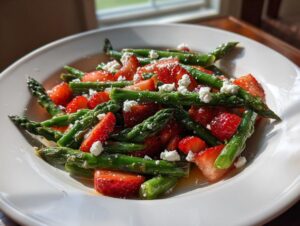Nahaufnahme eines Spargel-Erdbeer-Salat mit grünen Spargelstangen, roten Erdbeeren und zerbröseltem Feta auf einem weißen Teller.