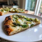 Nahaufnahme eines Stücks Spargel-Flammkuchen mit grünem Spargel und weißem Käse auf einem weißen Teller.