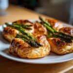 Mehrere goldbraune Päckchen Spargel im Blätterteig auf einem weißen Teller, frisch gebacken.