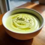 Nahaufnahme einer Schüssel mit cremiger, hellgrüner Spargelcremesuppe aus Spargelschalen, garniert mit Öl und Pfeffer.