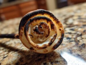 Nahaufnahme eines spiralig gewickelten Stockbrot vom Lagerfeuer, das leicht gebräunt und an den Rändern verkohlt ist.