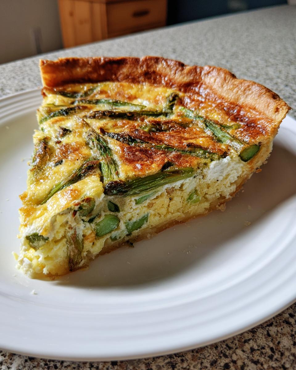 Nahaufnahme eines Stücks Knusprige Spargelquiche mit grünem Spargel und goldbraun gebackenem Teigrand auf einem weißen Teller.