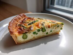Ein Stück cremige Spargel-Quiche mit grünem Spargel auf einem weißen Teller, im Sonnenlicht.