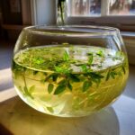 Nahaufnahme einer Glasschale mit Flüssigkeit und frischen Waldmeisterzweigen für die Klassische Maibowle.