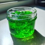 Nahaufnahme von leuchtend grünem Waldmeistergelee mit Apfelsaft in einem kleinen Glas, das im Sonnenlicht glänzt.