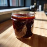 Dunkle, selbstgemachte Whiskey-BBQ-Sauce in einem kleinen Glas, das im Sonnenlicht auf einem Holzbrett steht.